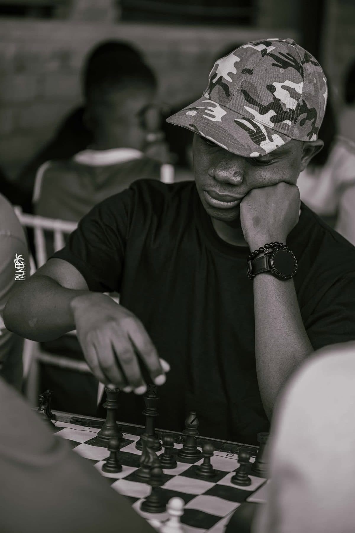 In black and white, camouflage cap, a hand moving a piece on the chessboard.